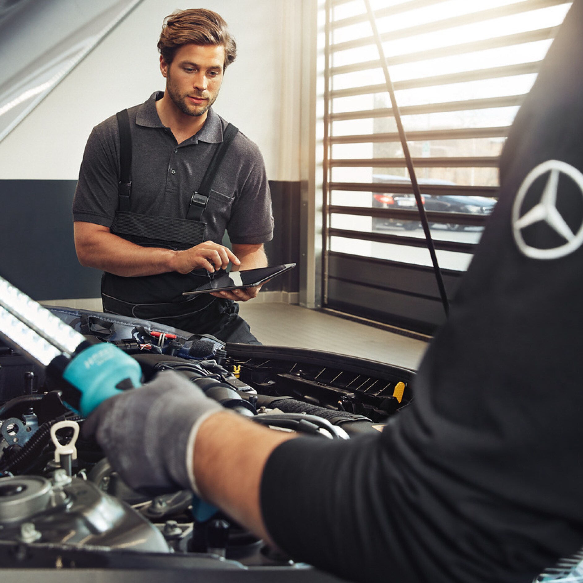 GenuineParts Two Mercedes-Benz technicians check the engine compartment of a vehicle.